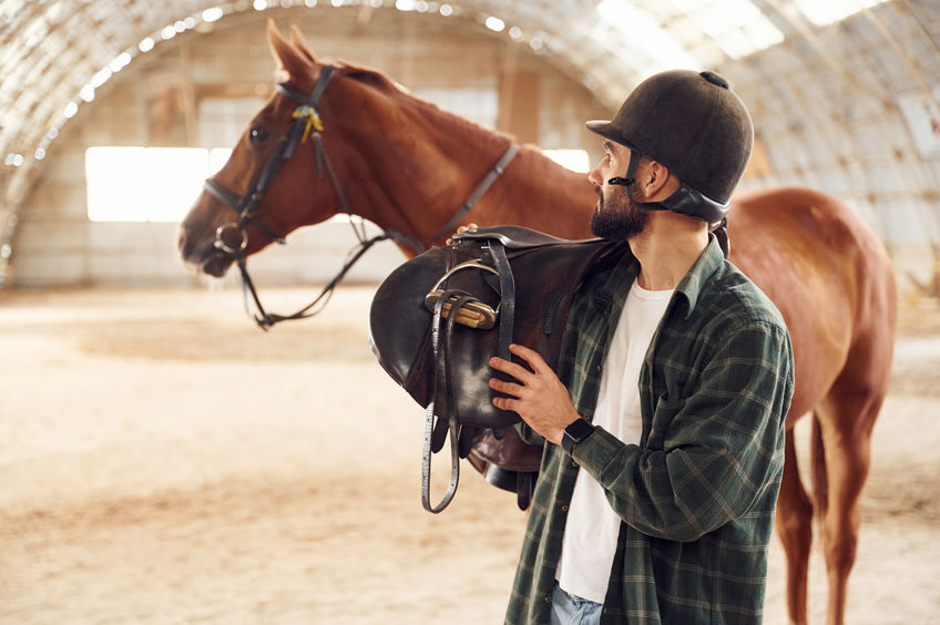 Holding the saddle in hands. Young man with a horse is in the hangar.